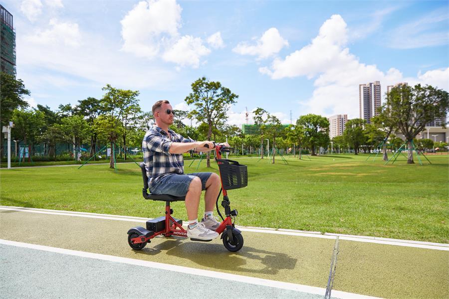 Man riding a red Hoverfly T3 three-wheel mobility scooter on a paved park path beside a grassy field
