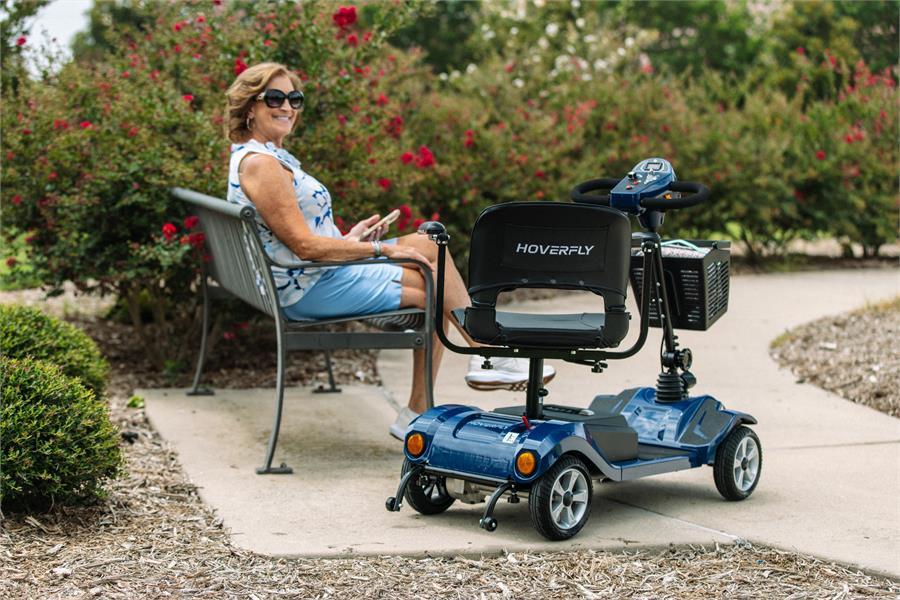 Older woman sitting beside a blue Hoverfly T4 mobility scooter on a garden path